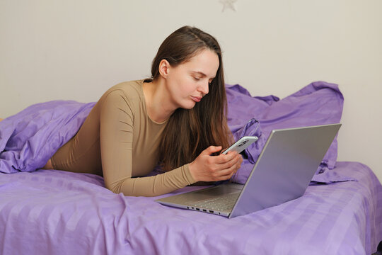 A woman is lying on a bed covered with purple blankets. She looks at her smartphone while a laptop is open beside her. Light fills the room from a nearby window