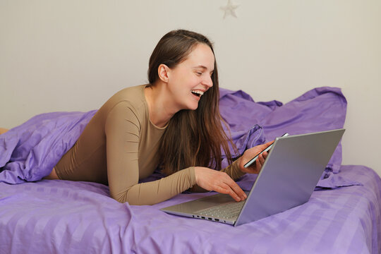 A woman is lying on her bed, using a laptop and holding a phone. She is laughing and appears to be enjoying her time. The room has a simple setting