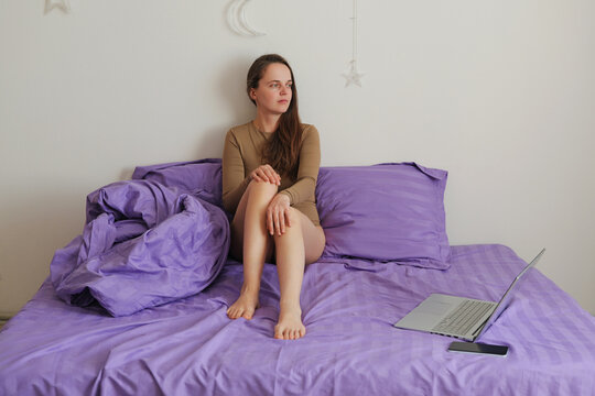 A woman sits on a purple bed in a simple room. She gazes out while a laptop is open beside her. The morning light fills the space softly