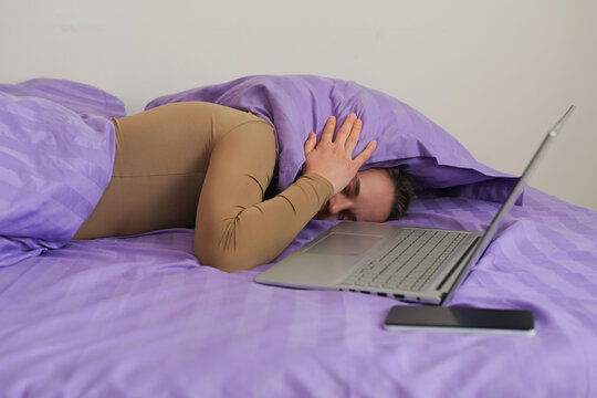 A person rests on a bed covered with purple sheets. They have a laptop and a phone placed next to them while they cover their head with the blanket