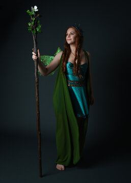 Full length portrait of female model wearing fantasy fairy costume, green dress, cloak, butterfly wings and crown. Full length walking pose holding druid staff.  isolated on dark studio background.