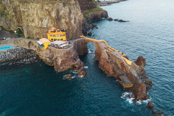 An aerial view showing a vibrant yellow restaurant perched on a rocky cliffside next to a historic stone bridge over the Atlantic Ocean in Madeira, Portugal. © JULIA