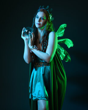 close up portrait of female model wearing fantasy fairy costume, green dress, butterfly wings and crown. standing pose holding  magical glass orb.  glowing green  lighting, dark studio background