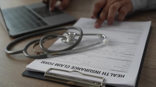 Doctor hand holds pen on health insurance claim form with stethoscope on clipboard, explaining medical policy details and healthcare coverage to patient on wooden office desk.