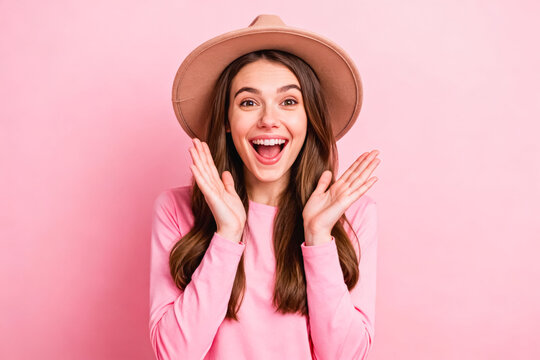 Studio portrait of a surprised excited young woman wearing a pink top and tan hat, clapping her hands with a joyful expression against a solid pink background.