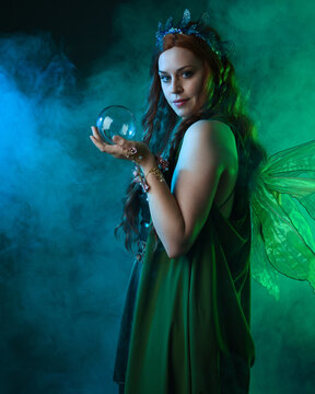 close up portrait of female model wearing fantasy fairy costume, green dress, butterfly wings and crown. standing pose holding  magical glass orb.  glowing green  lighting, dark studio background