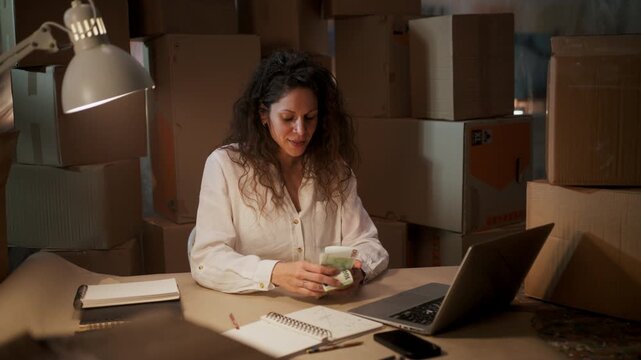 Successful female small business owner counting cash money at her desk in a warehouse.