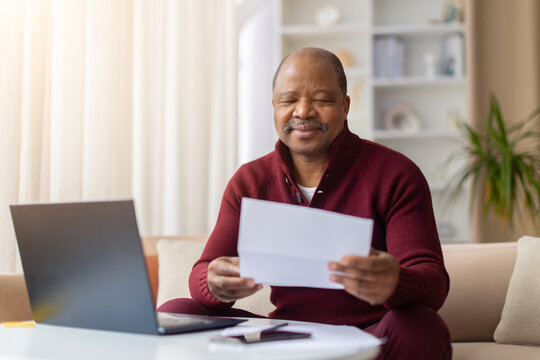 Pleased mature African American man reading paper document beside laptop at home, smiling contentedly. Financial paperwork, mail, or home office planning lifestyle concept indoors