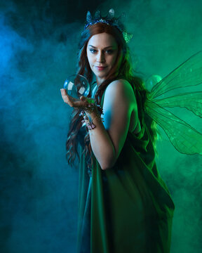 close up portrait of female model wearing fantasy fairy costume, green dress, butterfly wings and crown. standing pose holding  magical glass orb.  glowing green  lighting, dark studio background