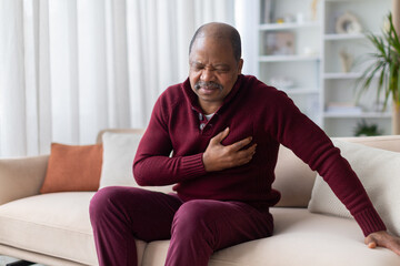 Fototapeta premium Mature African American man clutching his chest in pain while sitting on sofa at home. Heart attack, cardiac event, or chest pain medical emergency concept indoors