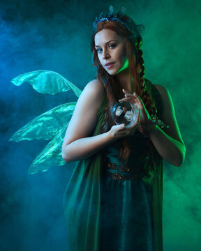 close up portrait of female model wearing fantasy fairy costume, green dress, butterfly wings and crown. standing pose holding  magical glass orb.  glowing green  lighting, dark studio background