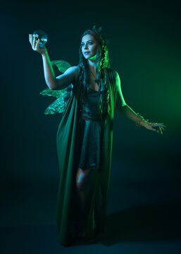 portrait of female model wearing fantasy fairy costume, green dress, cloak, butterfly wings and crown. full length standing pose, holding  magical crystal orb.  isolated on dark studio background.