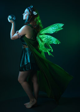 portrait of female model wearing fantasy fairy costume, green dress, cloak, butterfly wings and crown. full length standing pose, holding  magical crystal orb.  isolated on dark studio background.