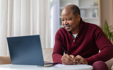 Smiling mature African American man taking notes while working on laptop at home desk. Remote work, studying, or financial planning lifestyle concept in living room interior © Anastasiya