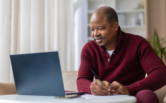 Smiling mature African American man taking notes while working on laptop at home desk. Remote work, studying, or financial planning lifestyle concept in living room interior