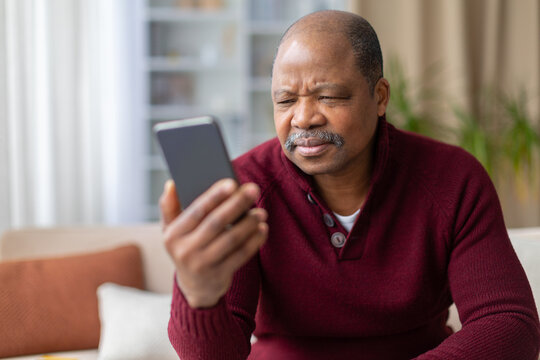 Confused mature African American man squinting and frowning at smartphone screen at home. Vision difficulty, bad news, or unclear content concept in domestic setting
