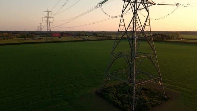 High angle aerial drone view close up of a steel lattice high voltage electricity pylon overlooking rural agricultural fields during a golden hour sunset near Doncaster UK