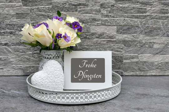 Blumenstrau&szlig; mit wei&szlig;en Rosen und lila Bl&uuml;ten in einer Vase neben einem Herz und Fotorahmen mit dem Text Frohe Pfingsten auf einem rundem Tablett vor einer grauen Ziegelwand.