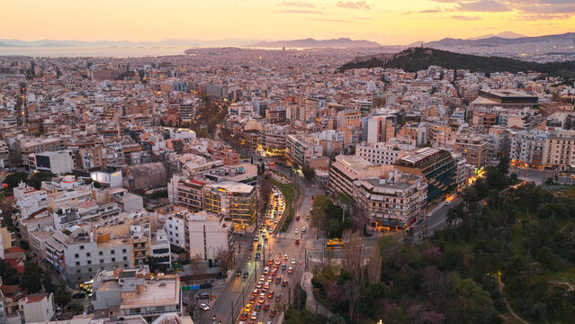Elevated view of athens at sunset, dense urban sprawl stretching to the sea and mountains, warm golden light, illuminated streets and flowing evening traffic