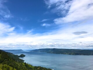 Fototapeta premium Azure sky drifts above calm waters. Green hills frame the distant shoreline. Lone boat glides on the tranquil surface. Sunlight dances softly across the lake's surface. Ideal for travel, nature