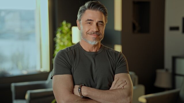 Portrait of a relaxed, smiling mature adult male with arms crossed in a stylish interior. Happy confident fit muscular mid adult older male in 50s with friendly smile. .
