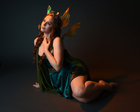 portrait of female model wearing fantasy fairy costume, green dress, cloak, butterfly wings and crown. full length sitting pose, kneeling on floor, dark studio background.