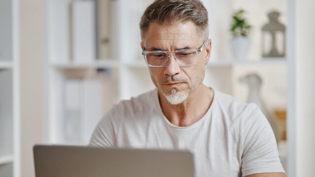 Middle aged casual man sitting at desk, looking at laptop, concentrating. Entrepreneur working from comfort of home office. Businessman listening on video meeting, making business decision, thinking.