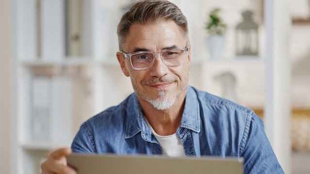 Portrait of middle aged man sitting at desk in bright room working with laptop computer in home office. Mature age, middle age, mid adult casual man in 50s, confident happy smiling.
