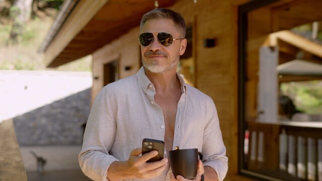 Businessman starting the day with morning coffee and checking phone. Portrait of middle aged man in sunglasses and casual cloth outdoor on home balcony. Spring or summer. Happy satisfied smile.