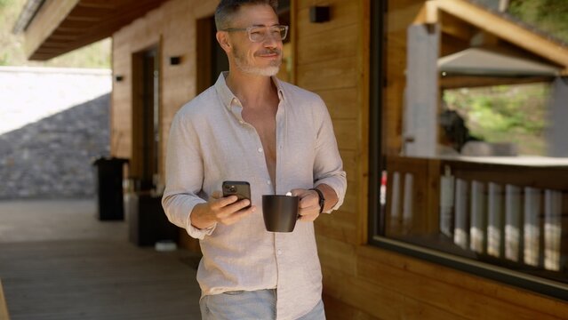 Casual middle aged man standing on balcony of wooden house, reading news on cell phone and drinking coffee. Perfect morning at home, happy satisfied smile.  .