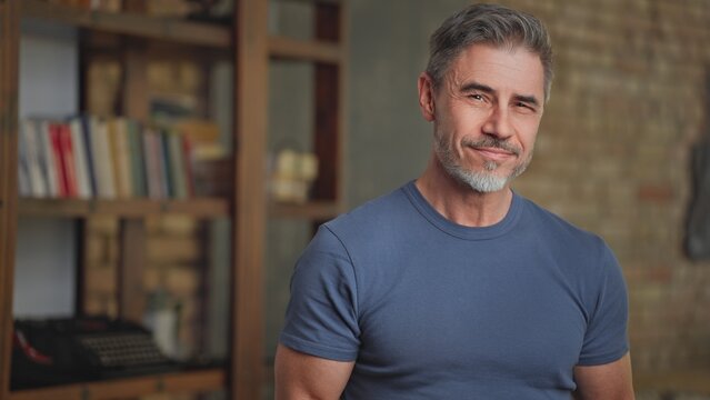 Portrait of happy confident casual middle aged man at home in loft room. Fit and healthy active senior male with grey hair looking at camera, smiling. Books in background, smart older guy.