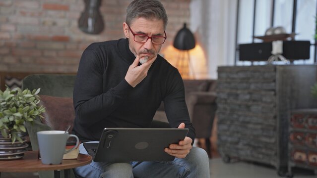 Thoughtful confident businessman working with laptop at home office desk. Middle aged man in glasses sitting in armchair, concentrating, browsing internet. Male entrepreneur remote managing business.
