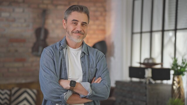 Portrait of happy casual middle aged man standing at home in loft living room. Senior male with grey hair in denim shirt looking at camera, smiling.