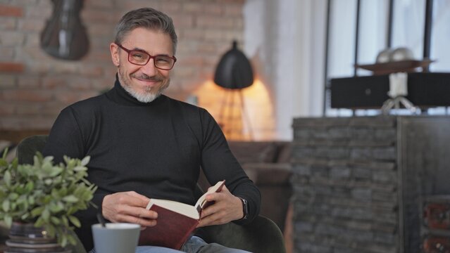 Smiling middle aged man sitting in living room reading book. Happy male wearing turtleneck pullover and eyeglasses. Smart looking older guy with grey hair enjoying leisure and free time at home.