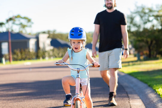 Happy young Australian kid riding her bike on quiet suburban road with parent following