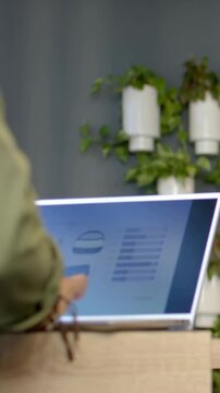 Vertical video: Typing olive-clad man at desk as peer hand pointing at laptop, plants, copy space