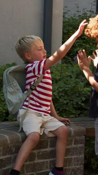 Vertical video: Reaching seated blonde student playing coming friend hands on brick ledge backpacks