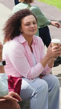 Camera framing seated woman in pink button shirt lifting paper coffee cup on bench, savoring sip