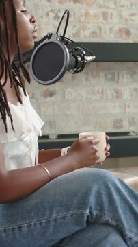 Vertical video: Woman sitting in studio leaning toward mic, starting podcast while sipping mug