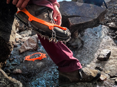 Close-up of a climber's hand and boot attaching orange ice crampons (anti-slip spikes) on a glacier surface. Safety equipment.