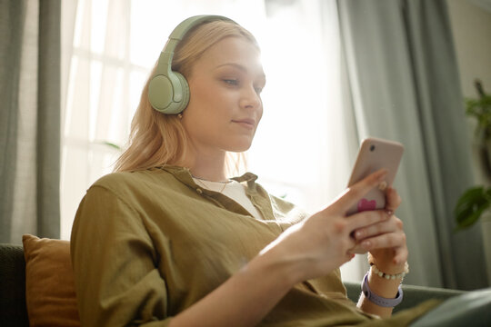 Young adult Caucasian woman wearing headphones and using smartphone for music streaming or mobile content consumption at home. Ideal for digital lifestyle, app promotion, audio services