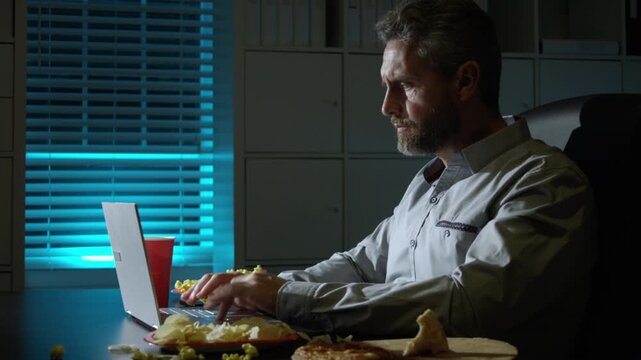 Late night work with laptop and snacks. Night scene in office. Late night work at desk eating chips. Office night snack break moment.