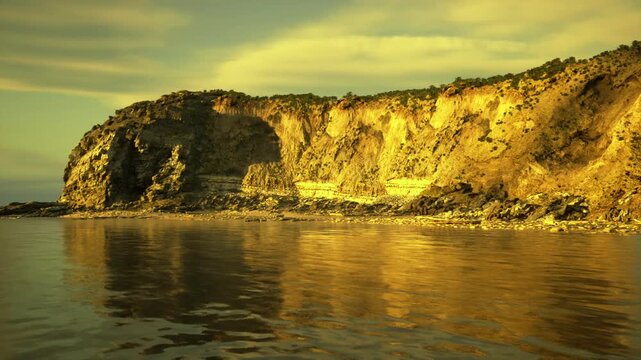 textured layered cliffs glowing in golden hour, coastal erosion and stratification clearly visible, shallow reflective water and scattered boulders provide