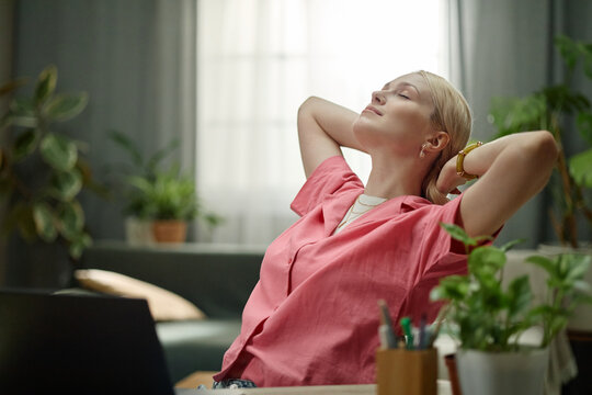 Caucasian young adult woman leaning back and stretching at desk beside laptop, taking break during home office work. Ideal for remote work, stress relief, productivity, wellness marketing