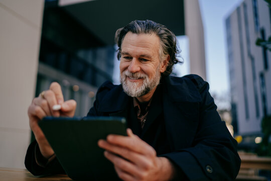 Happy senior professional using digital tablet with stylus, cheerful mature man working on creative project at outdoor cafe, digital nomad lifestyle, authentic representation, career growth.
