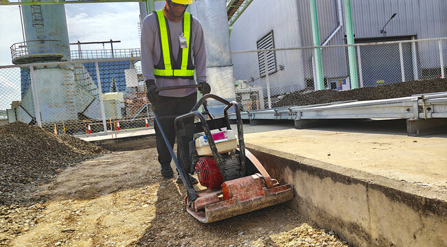 Worker compacting soil with tamping rammer near factory.