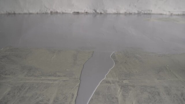 Closeup of worker applying gray epoxy resin paint to concrete floor. Roller spreads self leveling compound, creating smooth, durable, protective surface. Industrial renovation construction concept