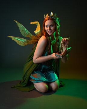 portrait of female model wearing fantasy fairy costume, green dress, cloak, butterfly wings and crown. Full length kneeling pose holding secret jewellery box, isolated on dark studio background