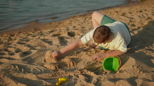 Preteen boy playing on sea shore with sand in warm sunset light. Child moving sand with hand. Development of sensory perception and creative creativity during natural interaction with nature.
