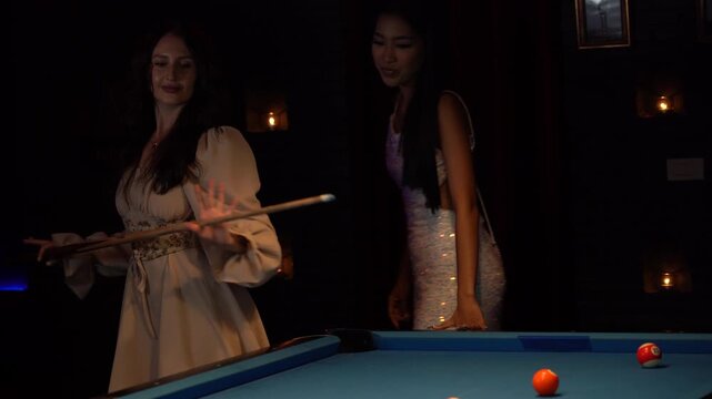 Beautiful woman playing billiards and dancing around the pool table with her friend. Young ladies having fun and celebrating in a nightclub with snooker. Nightlife, active leisure, and friendship.